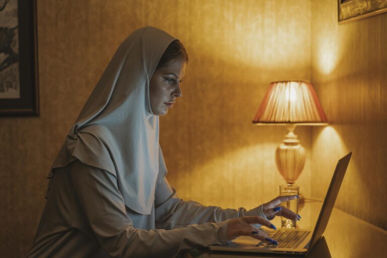 A woman in a hijab sits at a desk using a laptop, under warm lamplight.
